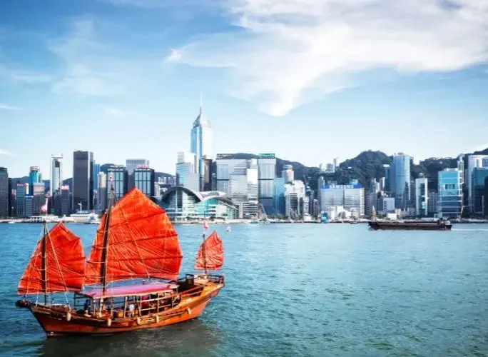 Traditional red-sailed Chinese junk boat crossing Victoria Harbour with Hong Kong's iconic skyline in the background
