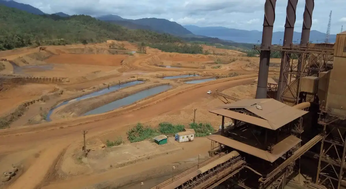 Aerial view of an open-pit mining site in Indonesia with industrial processing facility, settlement ponds, and forested mountains in the background
