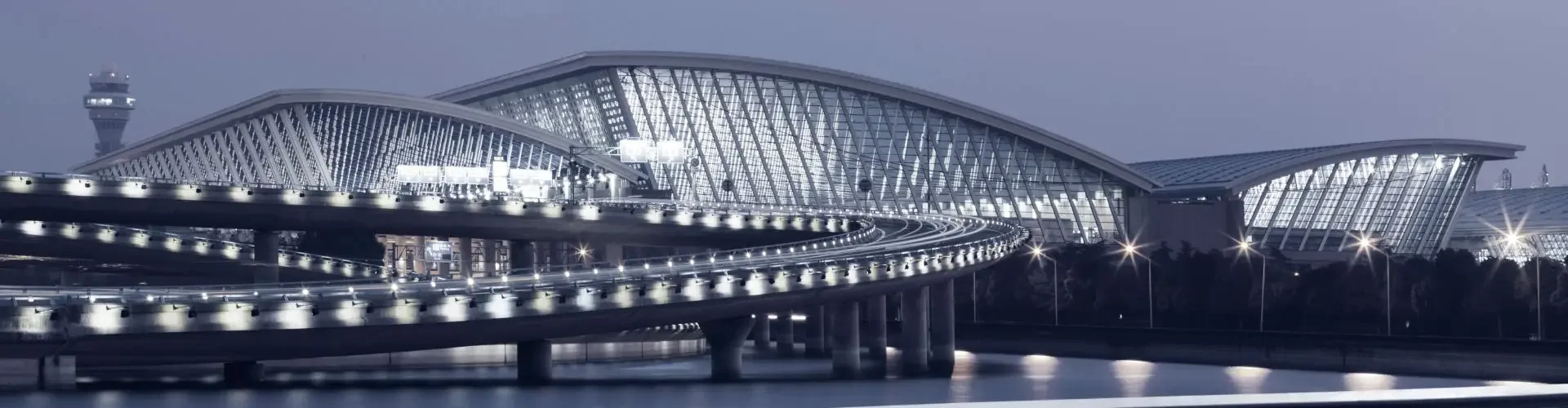 Shanghai Hongqiao Airport illuminated at dusk, showcasing its sweeping glass terminal and curved access road for private jet charter arrivals