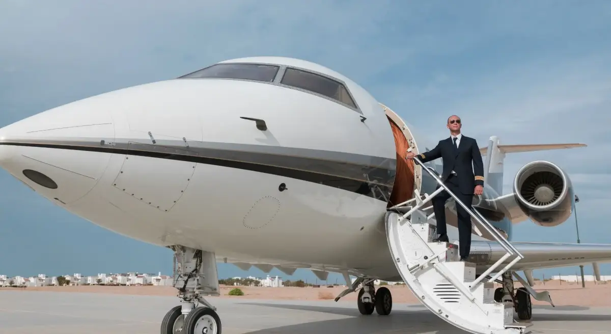Uniformed private jet pilot standing on aircraft stairs beside a business jet, ready to welcome corporate charter passengers