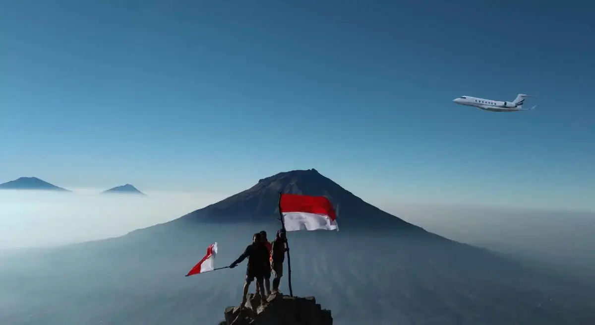 Two adventurers holding Indonesian flags on a volcanic mountain summit as a private jet flies overhead across the blue sky