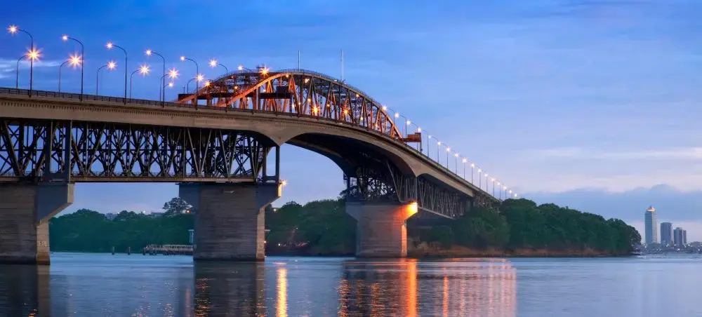 Auckland Harbour Bridge illuminated at twilight with city skyline reflecting in calm waters below