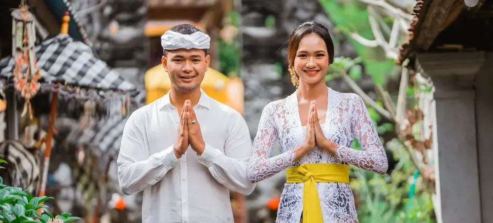 Balinese couple in traditional white ceremonial attire performing namaste greeting at ornate temple