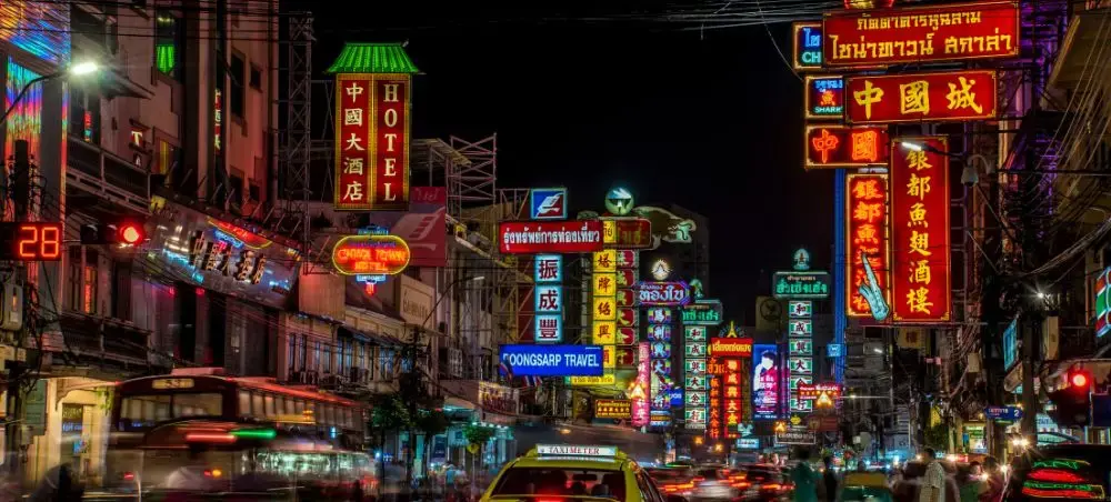 Vibrant neon signs illuminate bustling Bangkok street at night with colorful Chinese and Thai signage