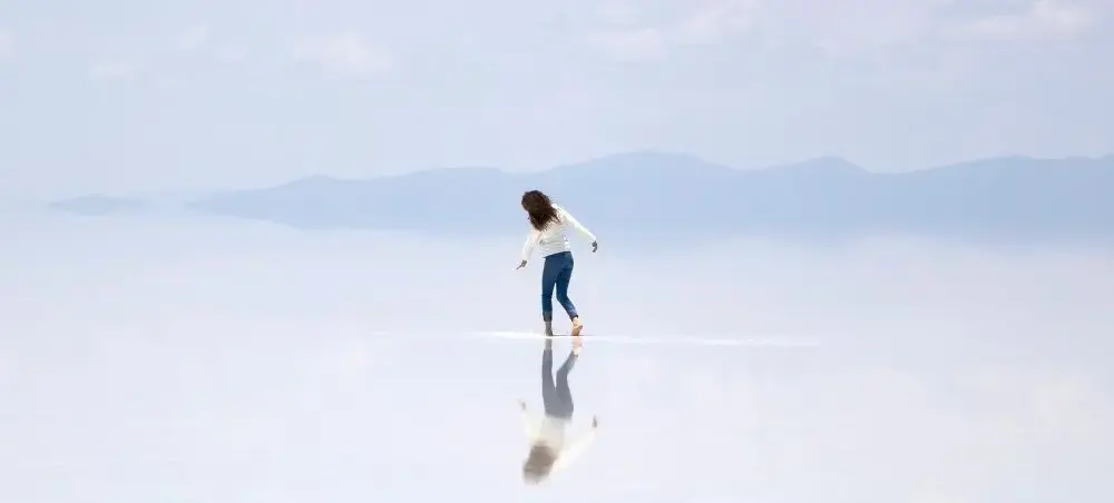 Woman walking across Bolivia's pristine Salar de Uyuni salt flats with mountain backdrop and reflection