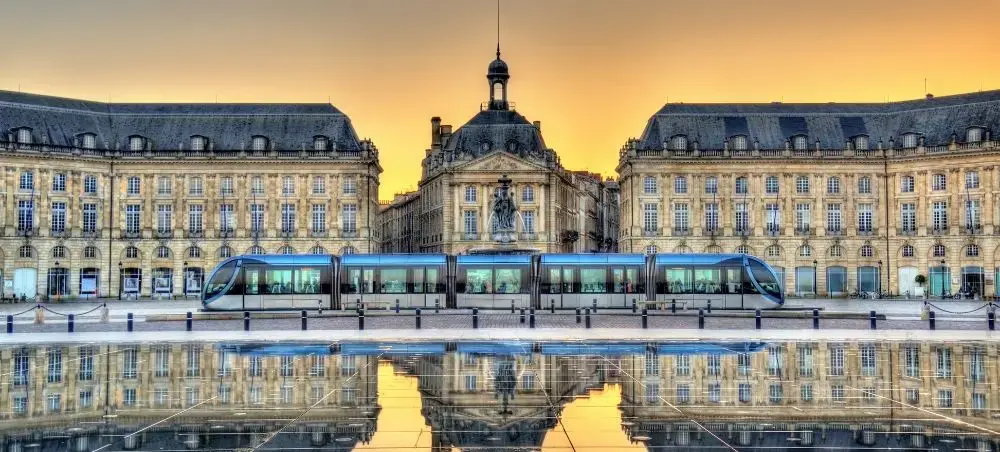 Modern blue tram gliding past historic Bordeaux architecture with golden sunset reflections on wet pavement