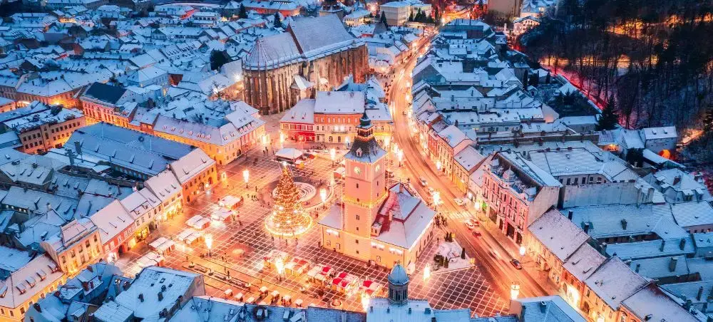 Aerial view of Brasov's illuminated medieval town square and historic buildings covered in winter snow at dusk