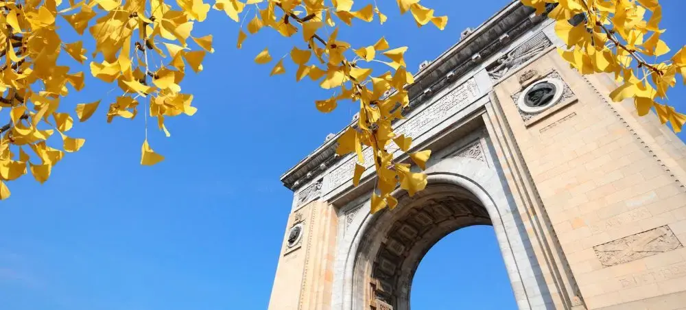Historic Bucharest triumphal arch framed by golden autumn leaves under brilliant blue sky