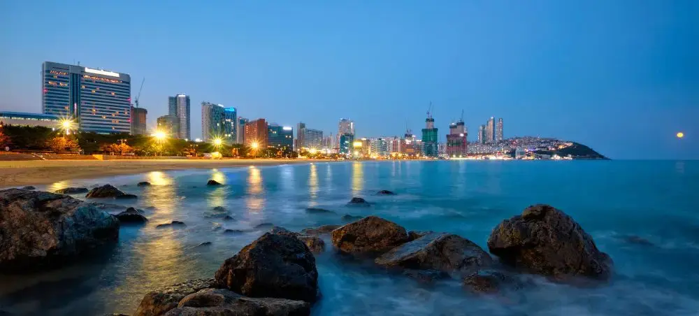Busan skyline illuminated at twilight with modern skyscrapers reflecting across calm ocean waters from rocky shoreline