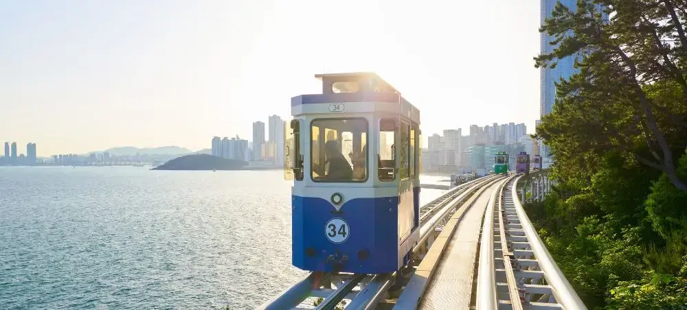 Blue vintage tram traveling along coastal railway tracks with Busan skyline and ocean views in background