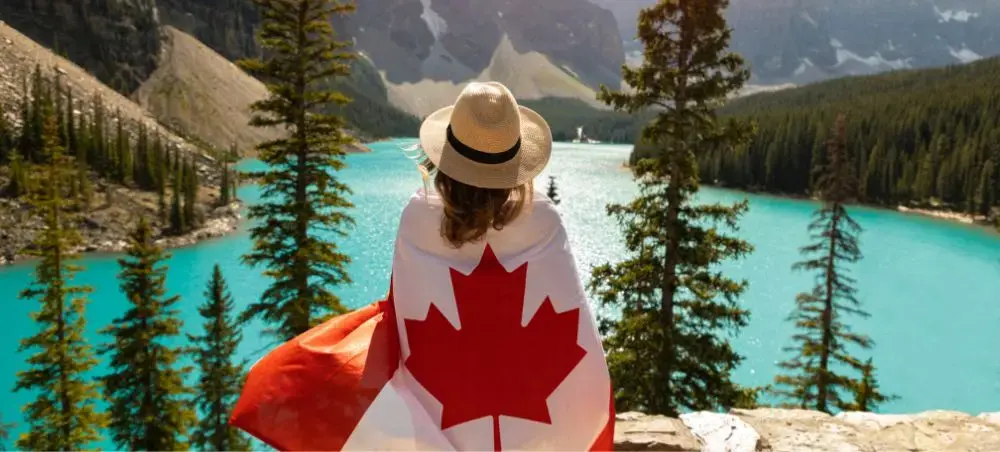 Traveler wrapped in Canadian flag overlooking turquoise mountain lake surrounded by evergreen forests