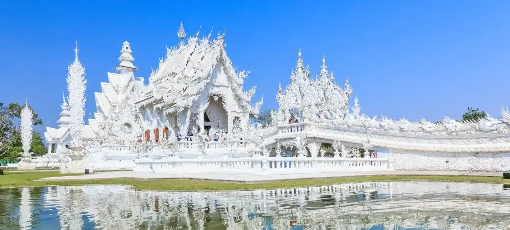 Stunning white Wat Rong Khun temple complex reflecting in tranquil waters under clear blue sky in Chiang Rai