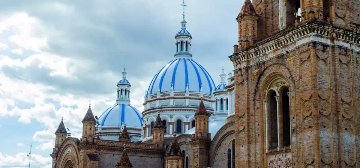 Historic blue-domed cathedral towers rise majestically above Cuenca's colonial architecture under dramatic cloudy skies