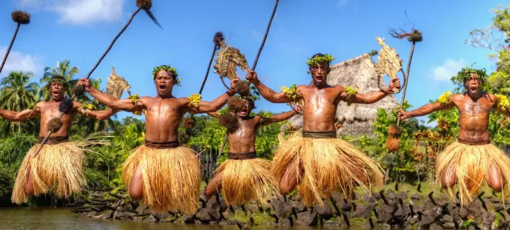 Traditional Fijian warriors performing ceremonial dance with spears and grass skirts in tropical village