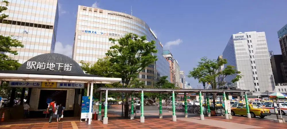 Modern Fukuoka city center with glass towers, underground shopping entrance, and bustling urban plaza