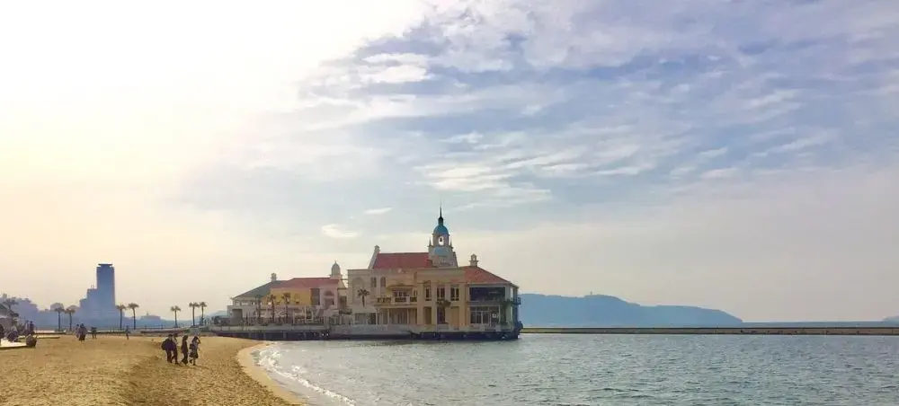 Historic waterfront building on stilts in Fukuoka Bay with sandy beach and mountain backdrop under cloudy sky
