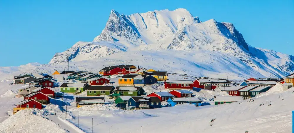 Colorful Arctic houses nestled in snow beneath towering Greenland mountains, showcasing remote luxury destinations