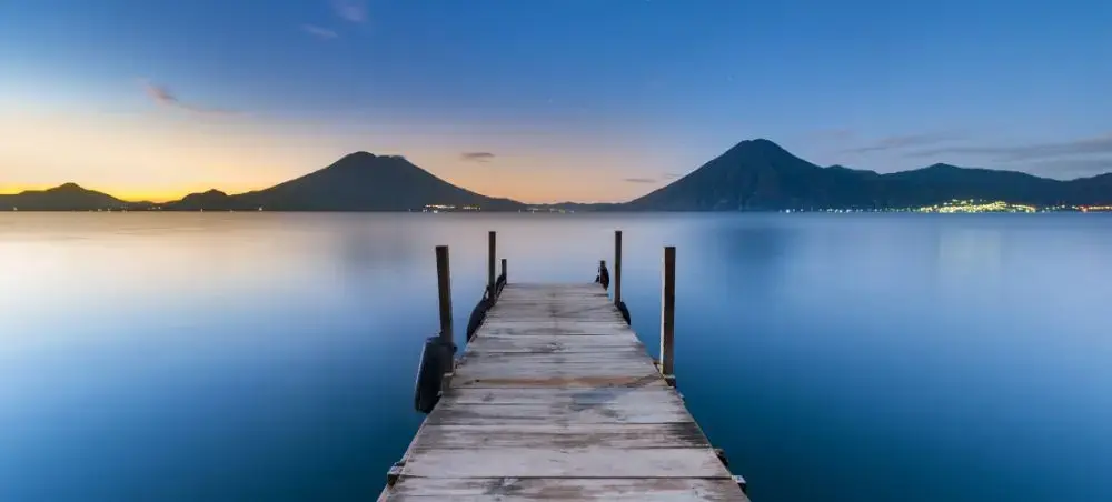 Wooden dock extending into calm lake waters at sunset with volcanic mountains silhouetted in Guatemala