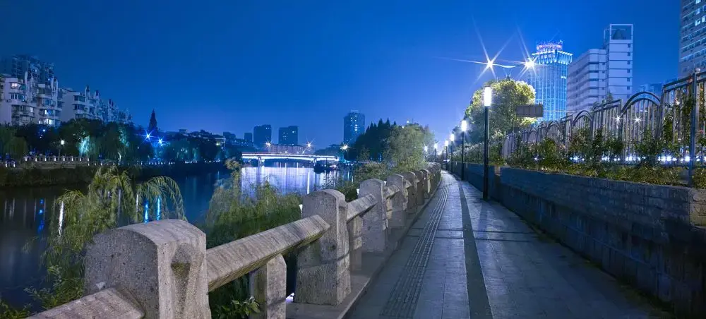 Illuminated stone walkway along Hangzhou's historic canal at twilight with modern city skyline and bridges