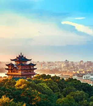 Traditional pagoda overlooking Hangzhou cityscape at sunset with lush green hills and modern skyline