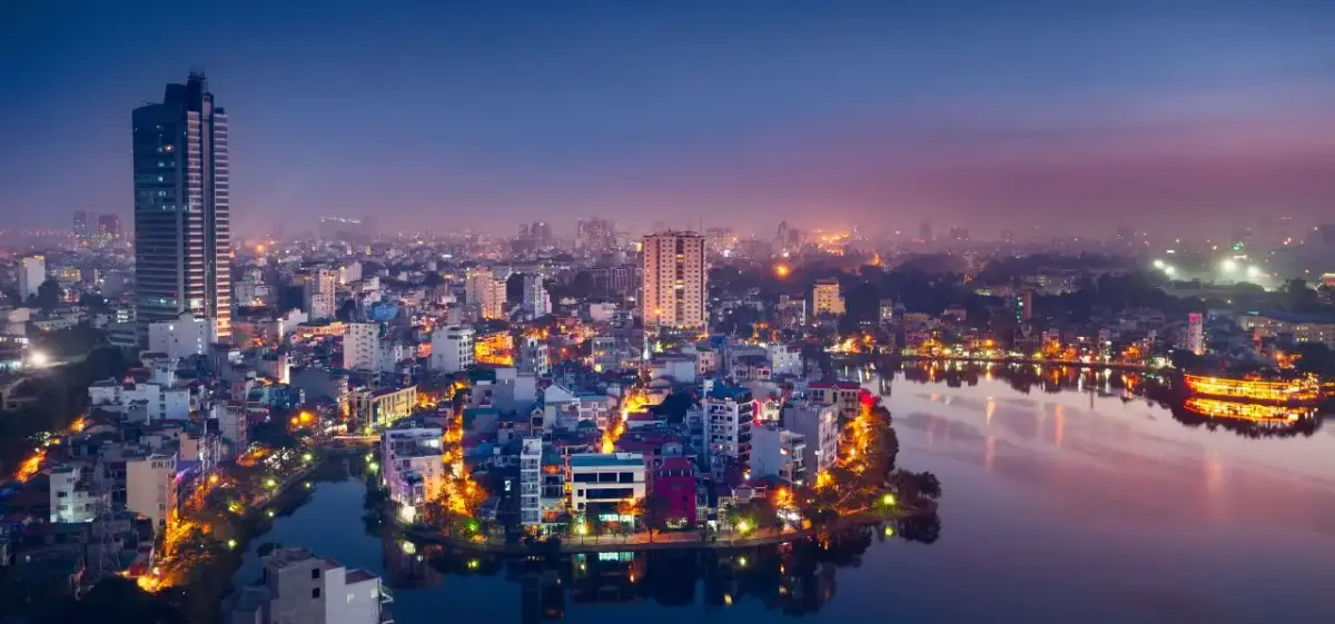 Hanoi cityscape at twilight with illuminated buildings reflecting in tranquil lake waters under purple sky