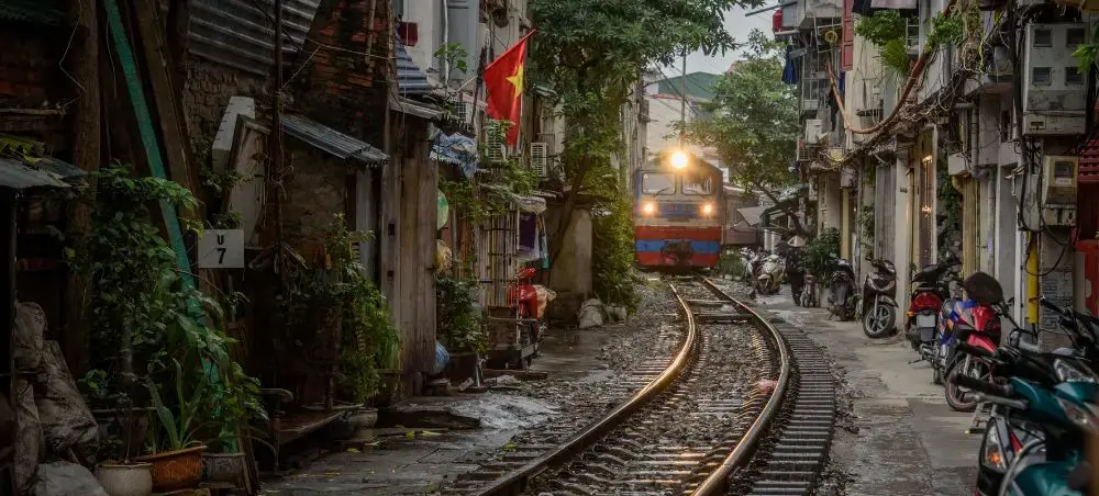 Train traveling through narrow residential street in Hanoi's Old Quarter with motorbikes and local homes