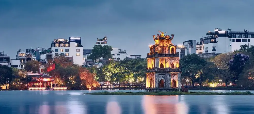 Illuminated Turtle Tower on Hoan Kiem Lake at dusk with Hanoi's modern skyline and traditional architecture