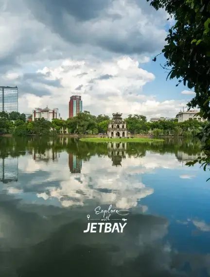 Historic Turtle Tower reflected in tranquil Hoan Kiem Lake waters with Hanoi cityscape and cloudy sky