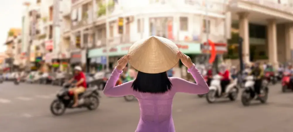 Woman in traditional Vietnamese ao dai and conical hat overlooking busy Ho Chi Minh City street scene