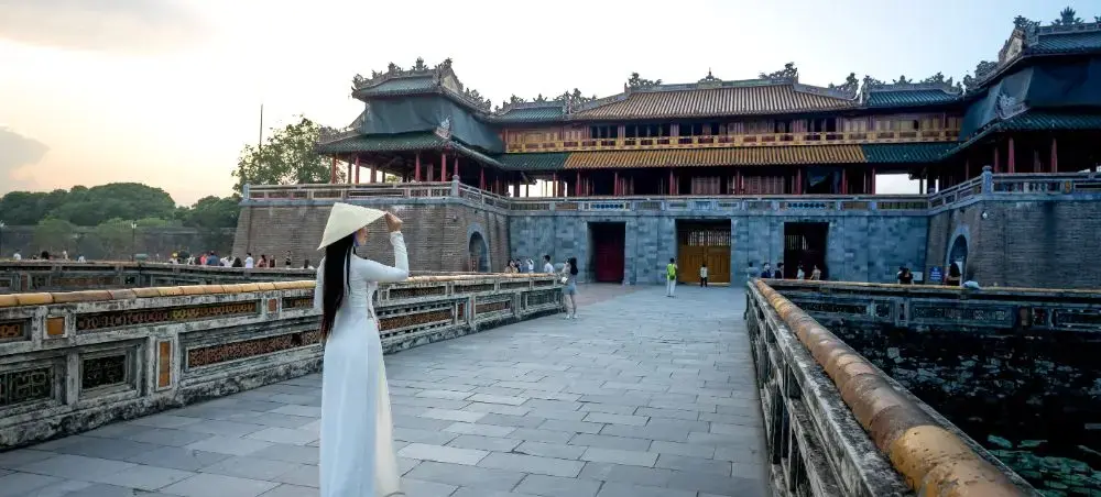 Woman in traditional Vietnamese ao dai walking across stone bridge at Imperial City of Hue during sunset