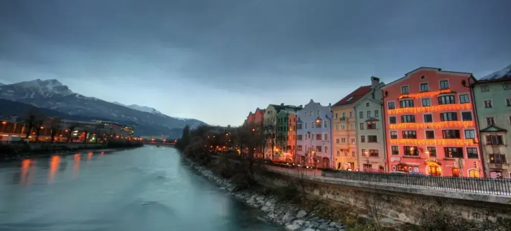 Colorful historic buildings along Inn River in Innsbruck at twilight with snow-capped Austrian Alps backdrop