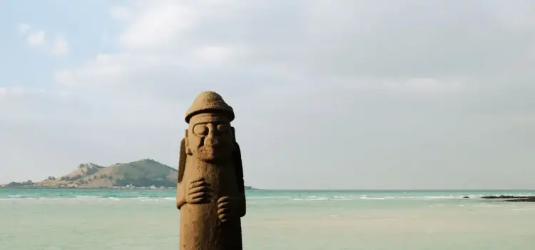 Traditional stone guardian statue on pristine Jeju Island beach with turquoise waters and volcanic hills