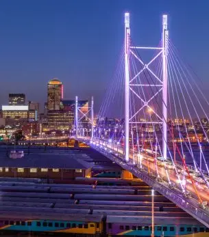 Illuminated Nelson Mandela Bridge spanning Johannesburg skyline at dusk with city lights and traffic below