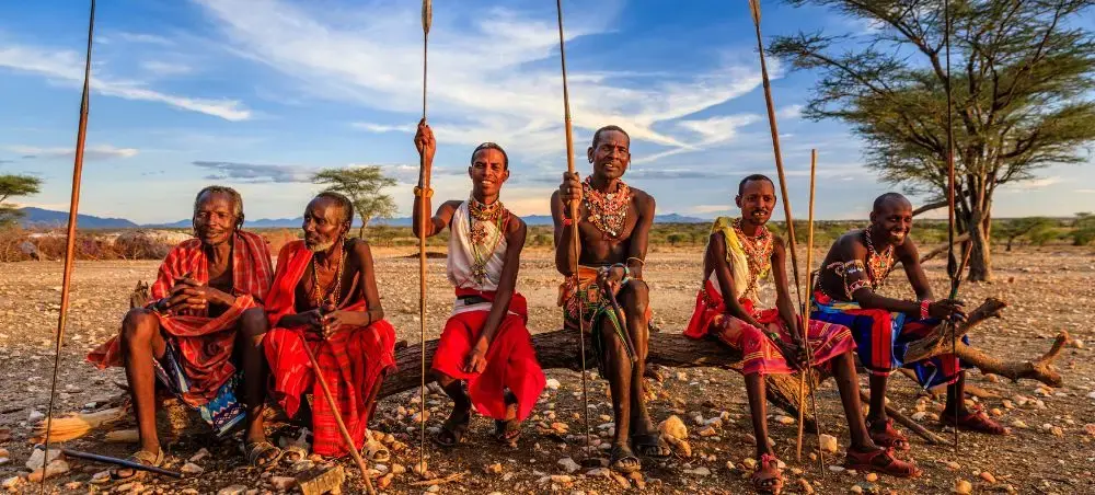 Maasai warriors in traditional red clothing and jewelry sitting with spears in Kenya's golden savanna landscape