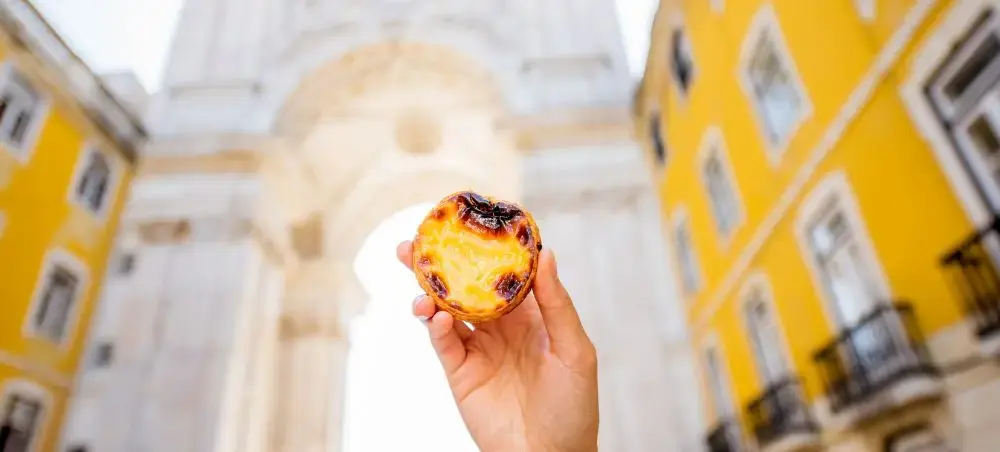 Hand holding traditional Portuguese pastéis de nata custard tart against Lisbon's historic yellow architecture