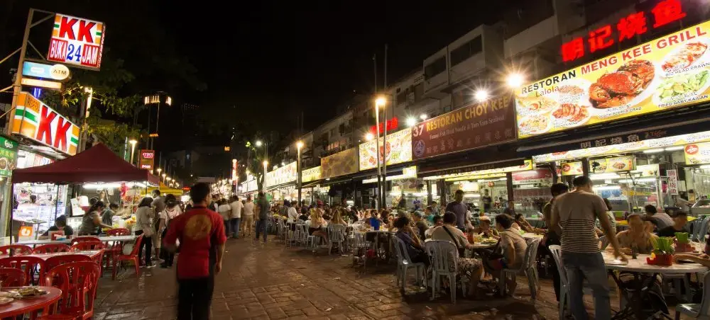 Bustling Malaysian street food market at night with diners enjoying local cuisine under bright neon signs