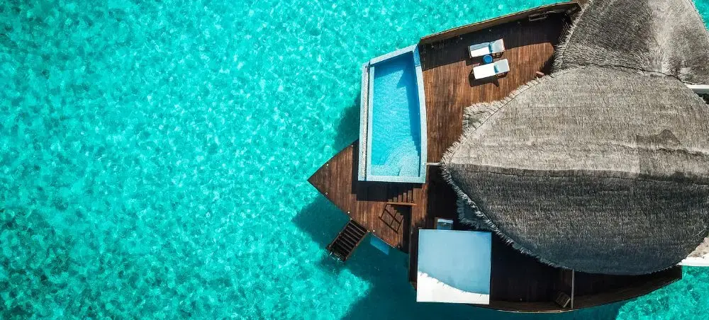 Aerial view of overwater villa with thatched roof and infinity pool in crystal clear Maldivian lagoon