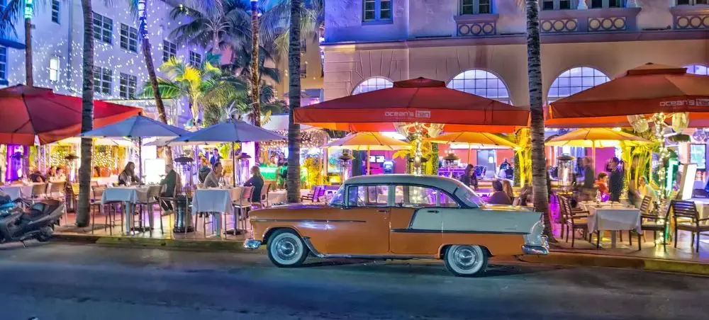 Vintage car parked outside vibrant Miami Beach restaurant with colorful umbrellas and palm trees at night