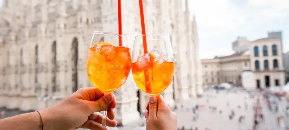 Two hands toasting with Aperol Spritz cocktails overlooking Milan's historic Duomo cathedral square