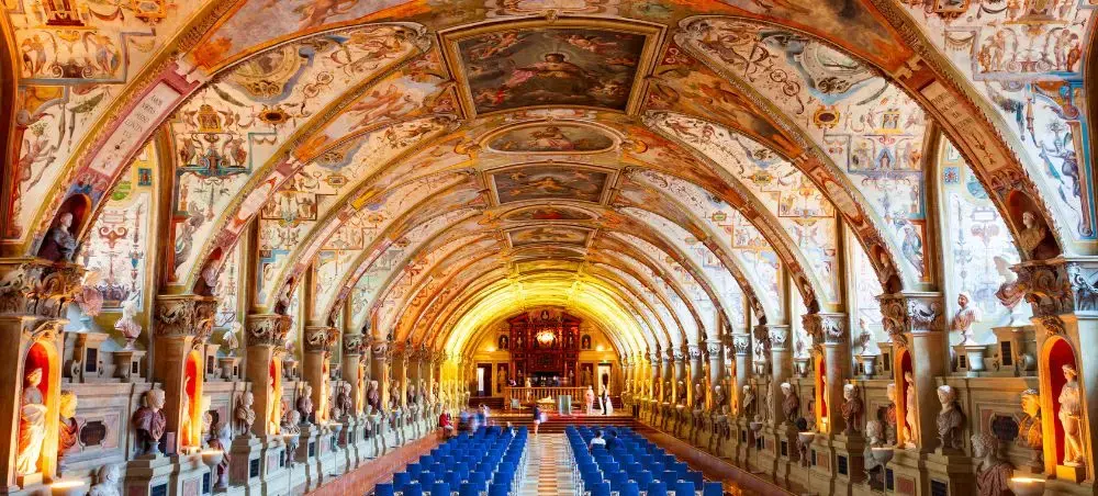 Ornate baroque hall with painted arched ceiling and blue chairs in Munich's historic Residenz palace