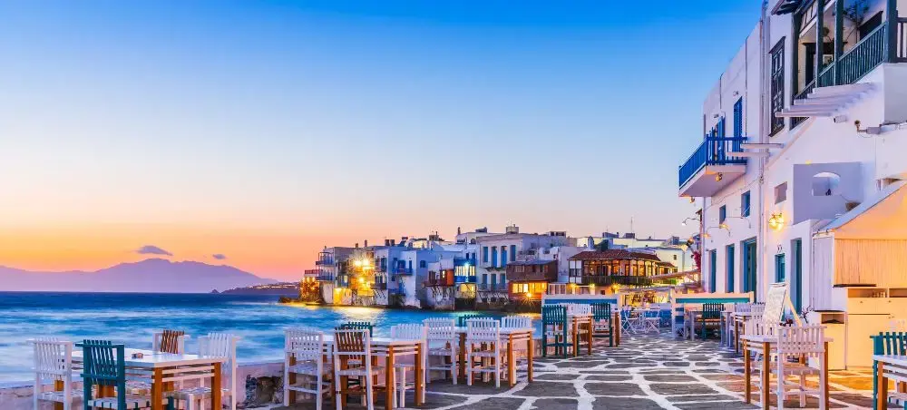 Sunset view of Mykonos waterfront with traditional white buildings, seaside restaurants, and golden hour lighting over the Aegean Sea