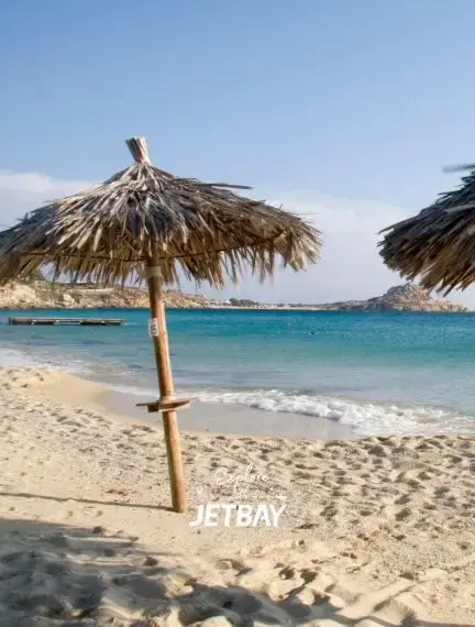 Traditional thatched beach umbrellas on pristine Mykonos shoreline with turquoise waters and luxury yacht