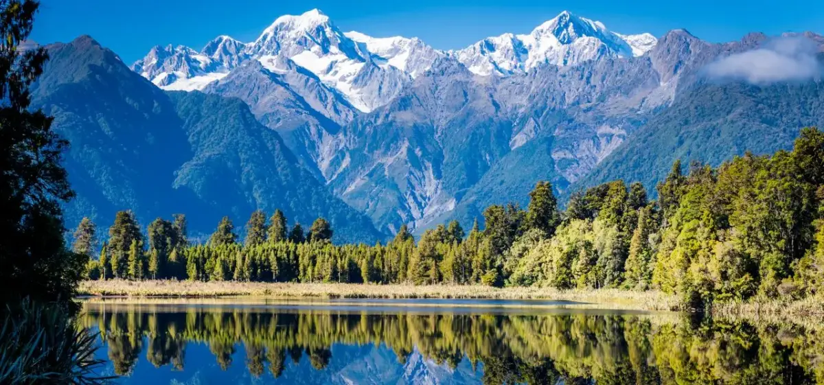 Snow-capped Southern Alps mountains reflecting in pristine lake surrounded by native New Zealand forest