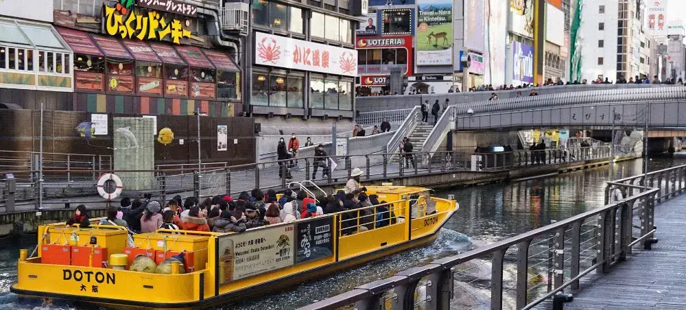 Yellow sightseeing boat filled with tourists navigating Osaka's Dotonbori River beneath neon signs and bridges