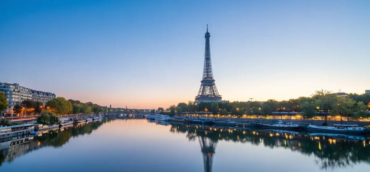 Eiffel Tower reflecting in Seine River at sunset with illuminated Parisian architecture and boats along waterfront