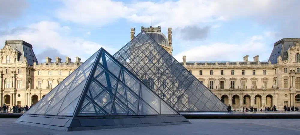 Glass pyramid entrance at the Louvre Museum in Paris with historic palace architecture under cloudy sky