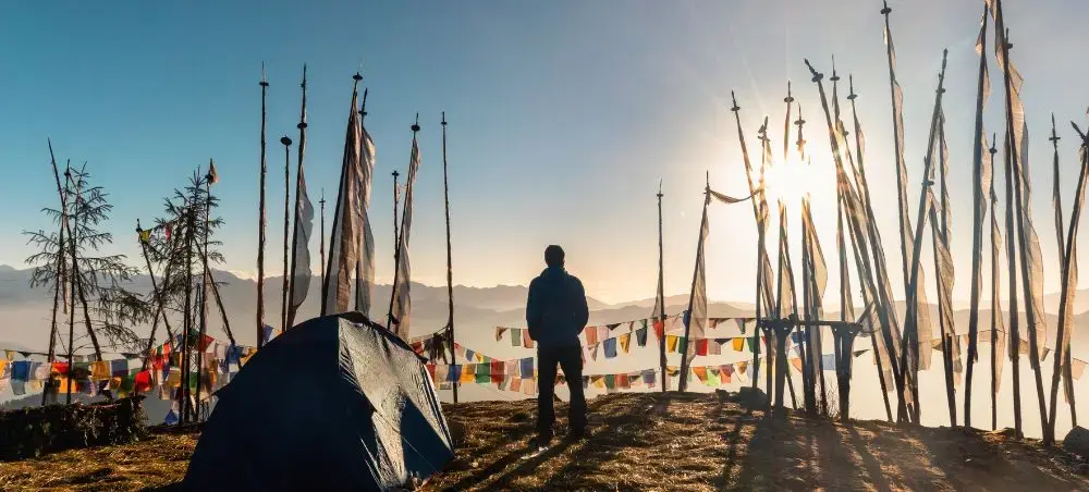 Traveler camping among colorful prayer flags at sunrise in Paro, Bhutan with mountain peaks backdrop