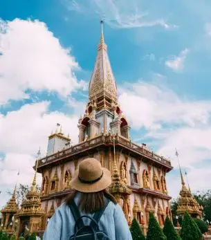 Traveler admiring ornate Buddhist temple spire in Phuket, Thailand under dramatic blue sky with clouds