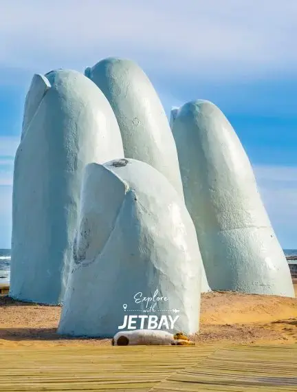 Iconic giant hand sculpture emerging from Punta del Este beach sand with Jetbay branding overlay