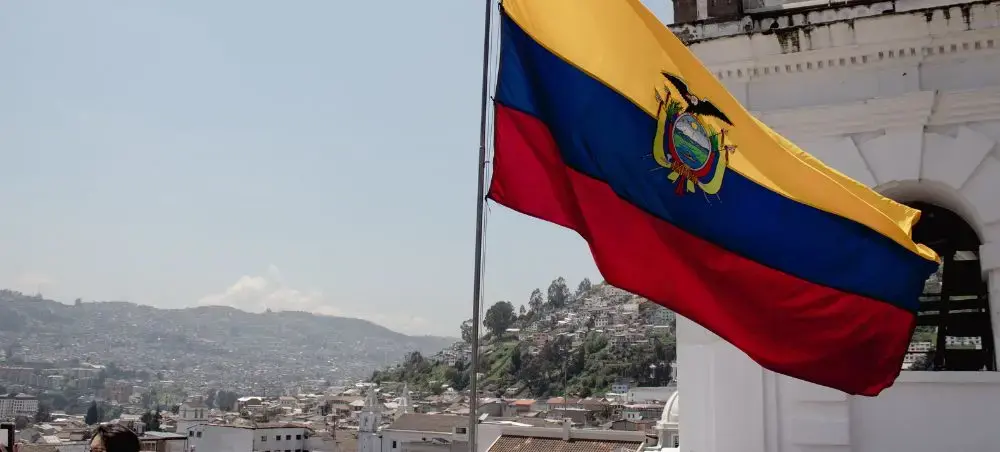 Ecuadorian flag with national coat of arms flying over Quito's hillside neighborhoods and mountains